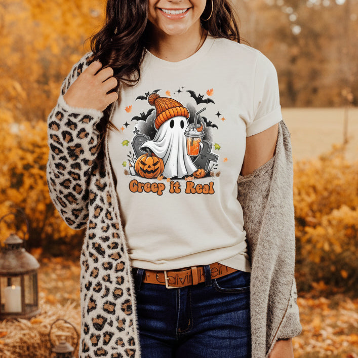 A smiling woman with dark curly hair stands in a seasonal, Fall background. She is wearing a cream-colored t-shirt with a cartoon graphic of a ghost in a beanie, holding an iced drink, and surrounded by bats and pumpkins with the text "Creep it Real." She is wearing a leopard print cardigan over the shirt, dark jeans, and a Calvin Klein belt.
