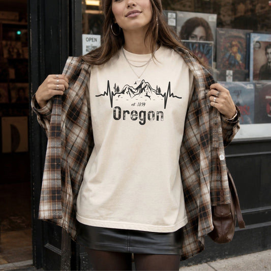 A woman wearing a cream graphic t-shirt with a black mountain and heartbeat design that reads 'Oregon', styled with an open brown plaid flannel shirt and a black leather skirt, standing in front of a storefront window.