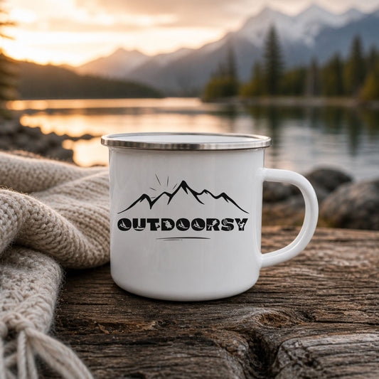 A white enamel camping mug with a silver rim and handle, featuring a black mountain logo and the word 'OUTDOORSY', set on a rustic wooden surface next to a knitted blanket with a blurred mountain lake background at sunset.