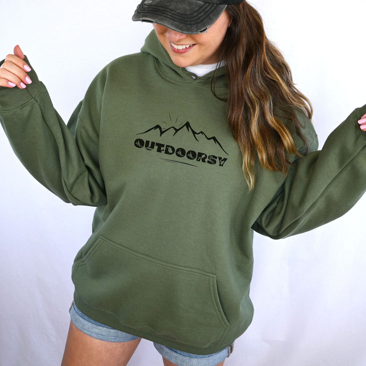 Woman outdoors wearing a Military Green hoodie with a minimalist mountain outline design and the word Outdoorsy featuring embedded adventure silhouettes.
