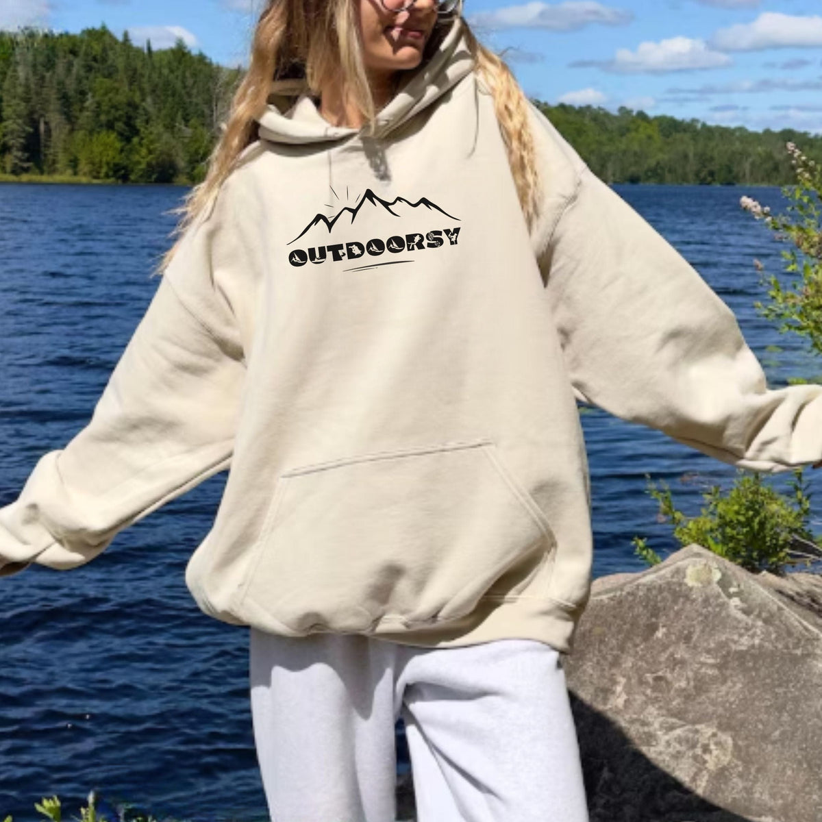 Person wearing a beige hoodie with 'OUTDOORSY' text by a lake