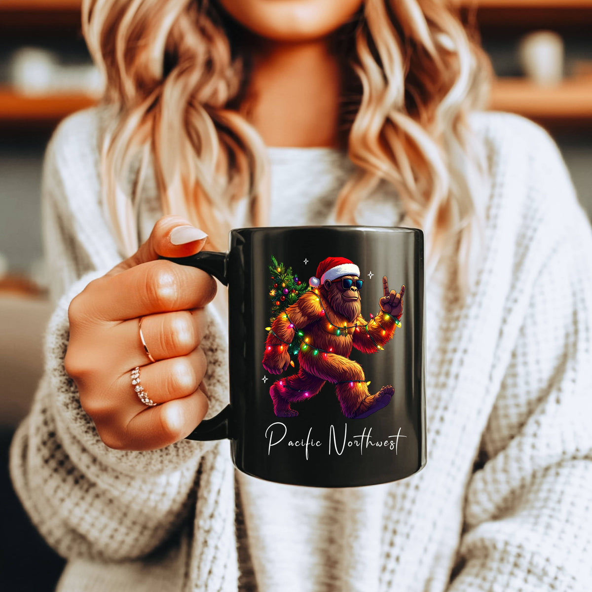 A woman in a white knit sweater holds a black ceramic mug featuring a colorful graphic of a Santa-clad bigfoot with the text 'Pacific Northwest' on a warm indoor background.