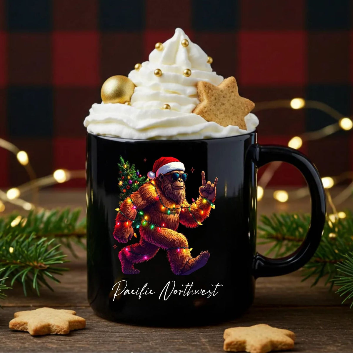 A black ceramic mug filled with whipped cream, gold sprinkles, a cookie, and a star cookie, set on a wooden surface with holiday lights, pine branches, and a red plaid backdrop. The mug features a graphic of Bigfoot in a Santa hat and jeans dancing with a Christmas tree, with the text 'Pacific Northwest' below.