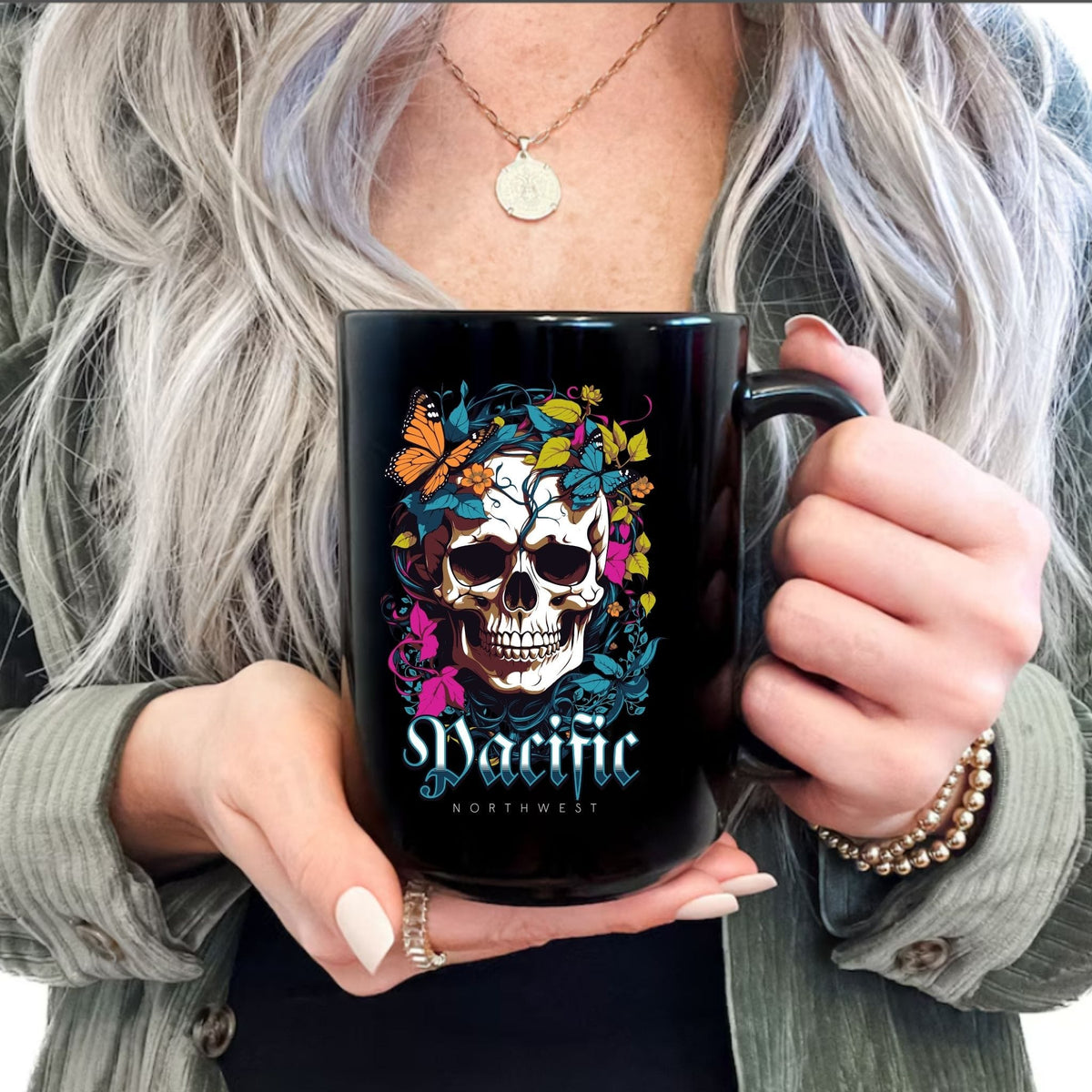 A person holding a black ceramic mug with a colorful skull graphic and the text 'Pacific Northwest'. The person has long silver-gray hair, wearing a green cardigan over a black top.