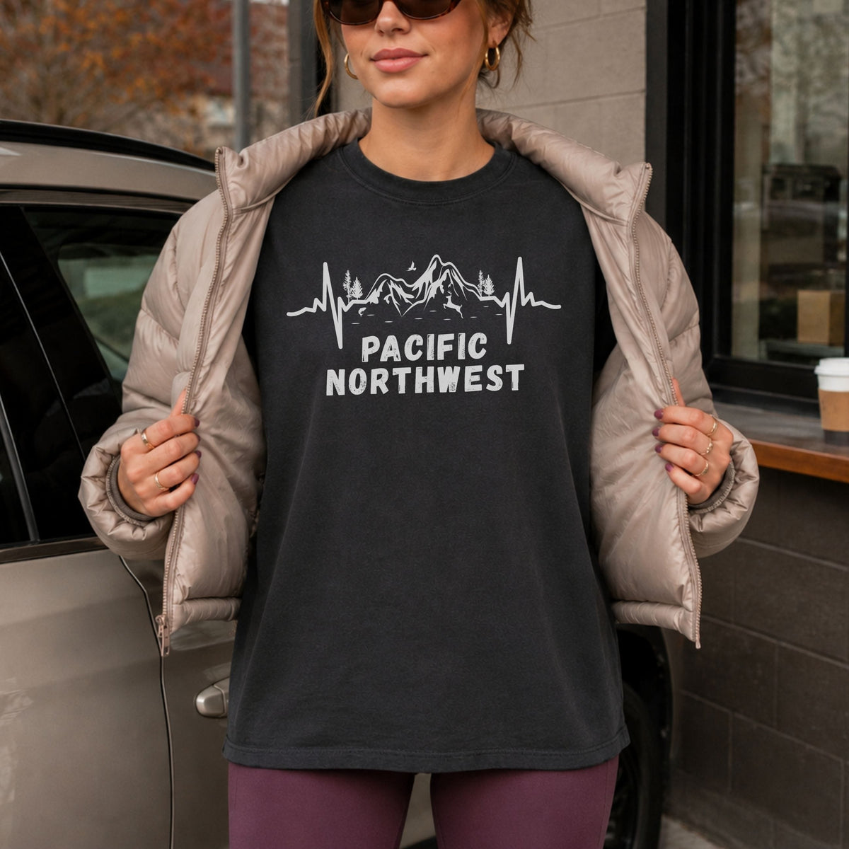 Woman wearing a black Pacific Northwest Comfort Colors T-shirt with a white mountain heartbeat graphic, layered under a taupe puffer jacket beside a parked car and café window.