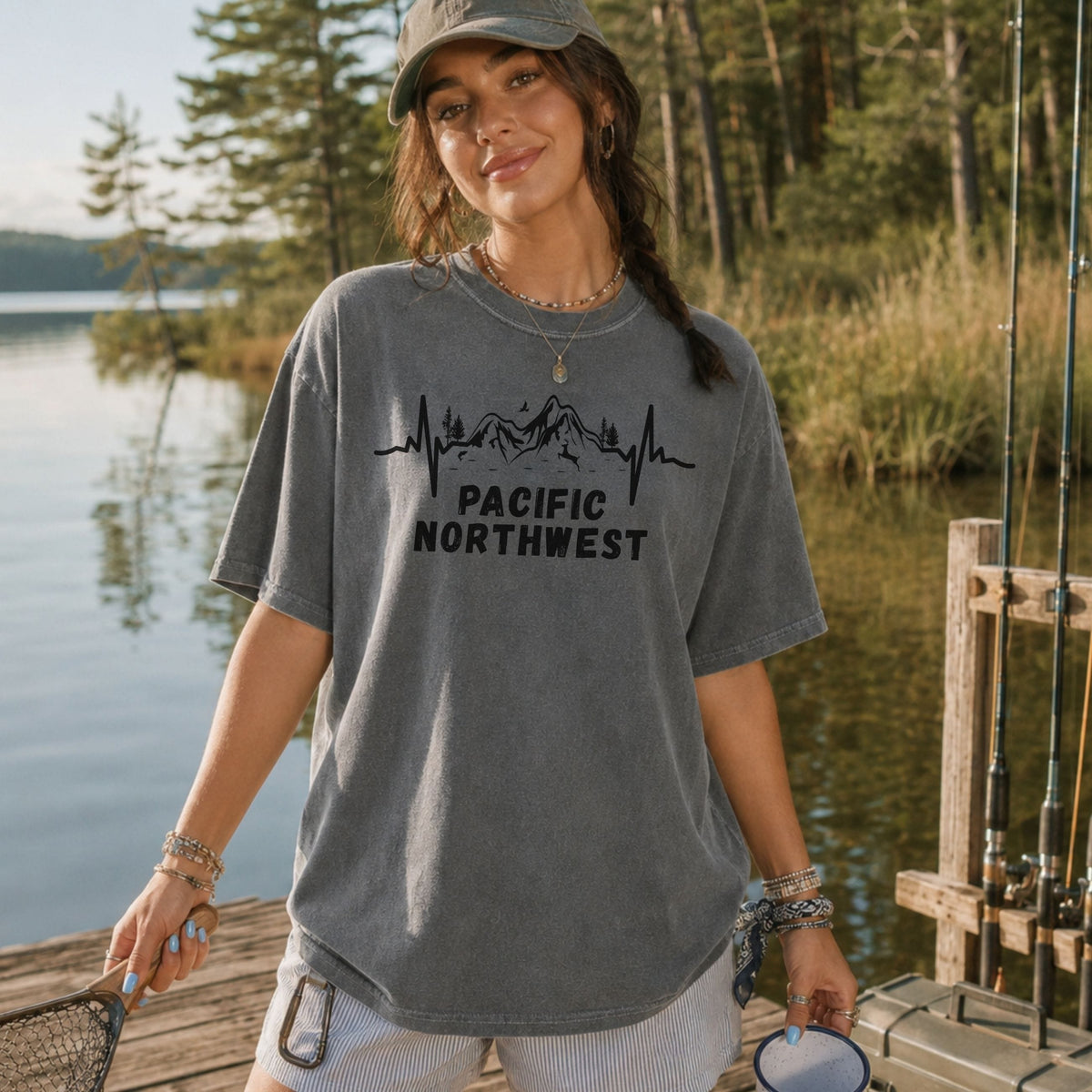 A woman wearing an oversized gray t-shirt with a black mountain and heartbeat graphic that reads 'PACIFIC NORTHWEST' stands on a wooden dock by a lake with trees in the background.