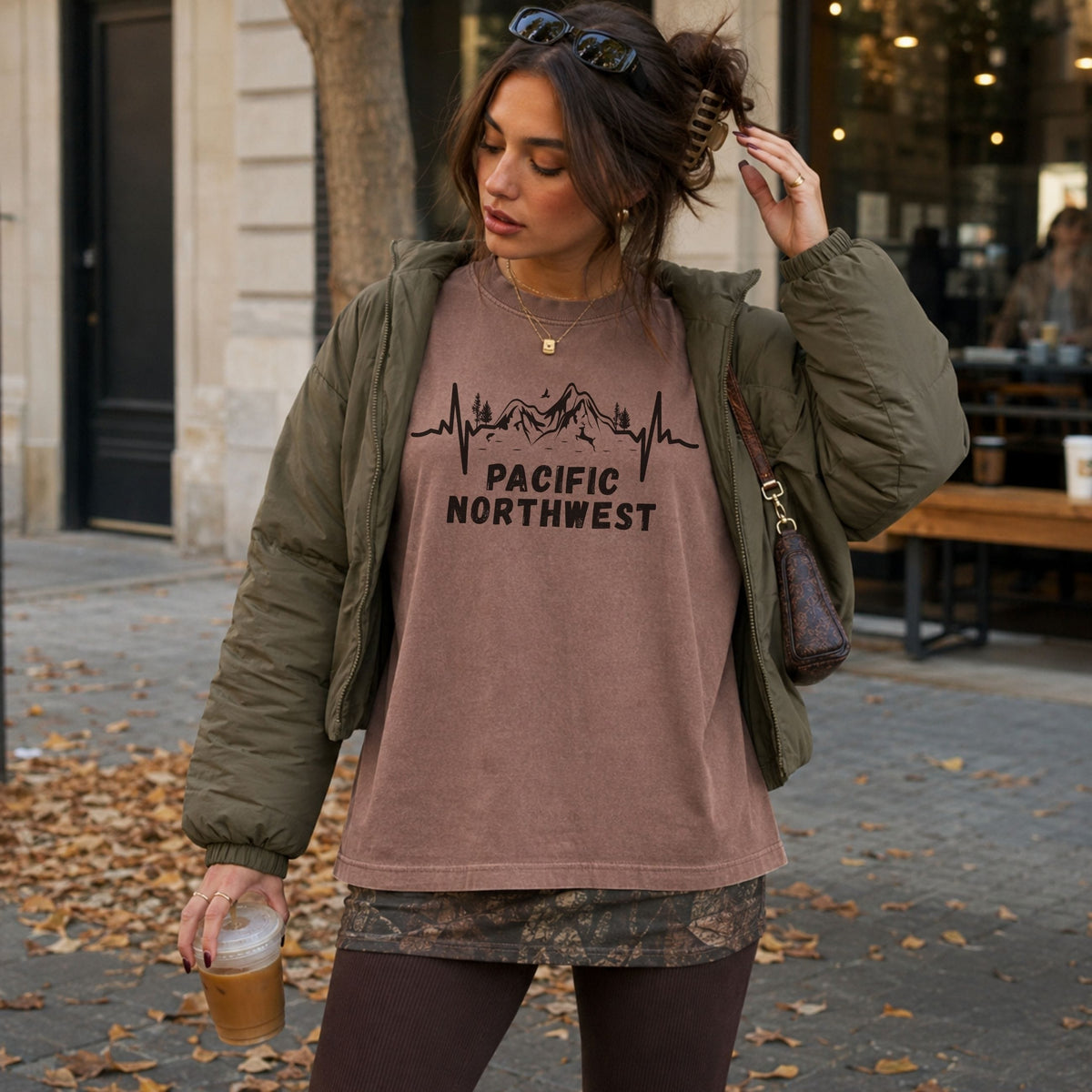 Woman wearing a brown Pacific Northwest Comfort Colors T-shirt with a black mountain heartbeat graphic, layered under an olive jacket while standing outside a café on a fall day.