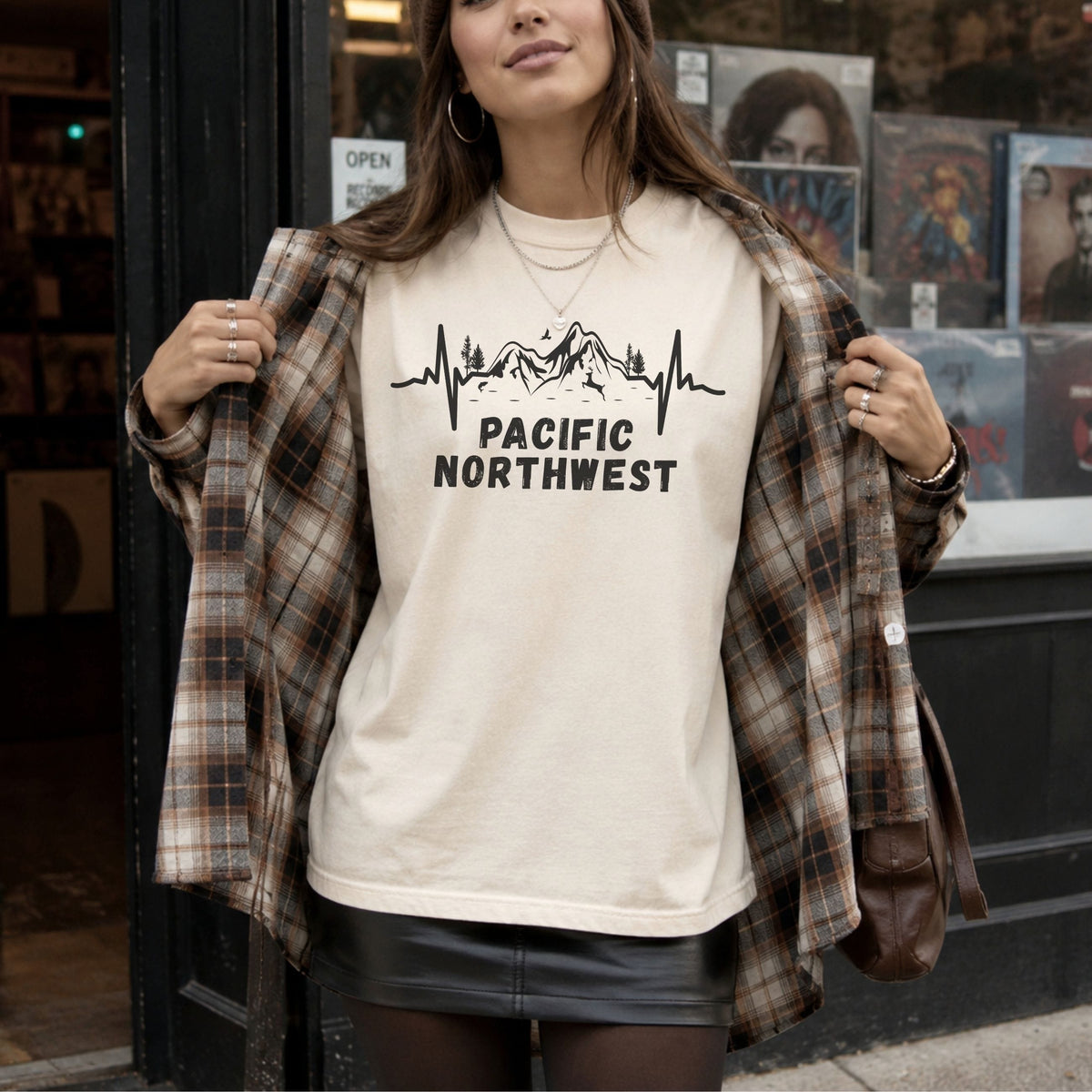 Woman wearing an ivory Pacific Northwest Comfort Colors T-shirt with a black mountain heartbeat graphic, layered under a brown plaid overshirt outside a record shop.