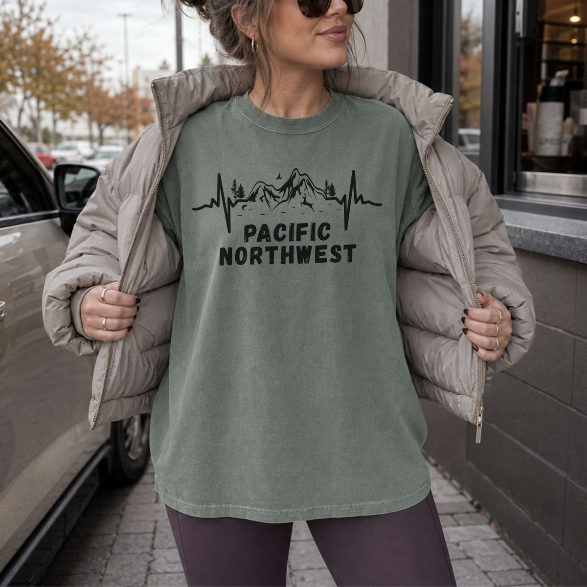 Woman wearing a moss green Pacific Northwest Comfort Colors T-shirt with a black mountain heartbeat graphic, layered under a puffer jacket outside a coffee shop.