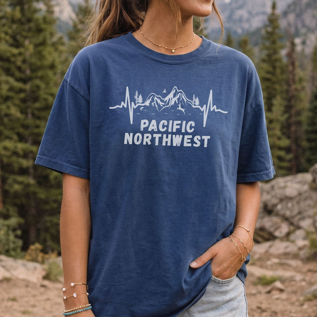 Woman wearing a navy blue Comfort Colors T-shirt with a white mountain heartbeat and 'Pacific Northwest' graphic, standing outdoors on a rocky mountain trail with evergreen trees in the background.