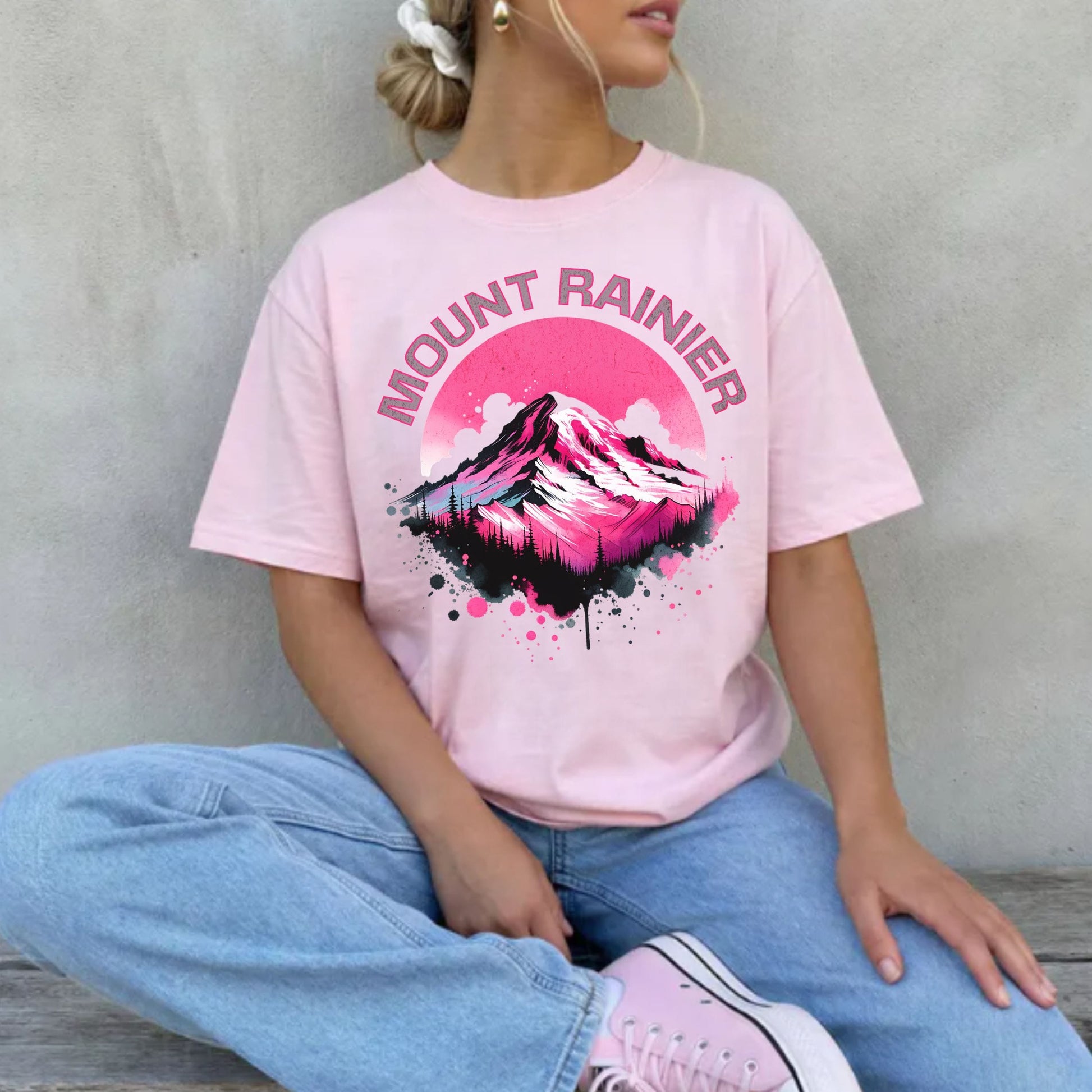 A woman wearing a light pink graphic t-shirt with a 'MOUNT RAINIER' mountain design in pink, black, and white, paired with light blue jeans and pink sneakers, sitting against a textured gray wall.