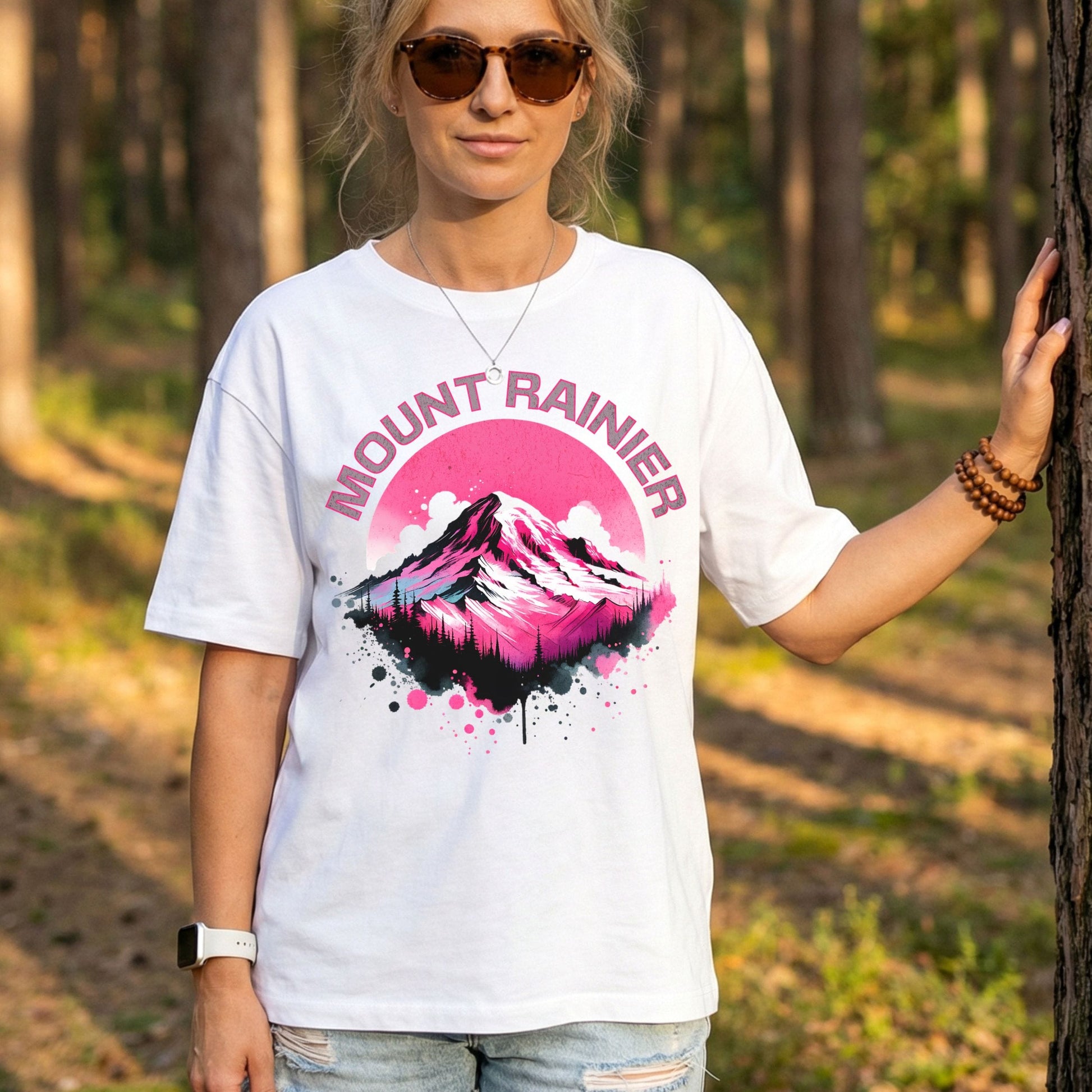 A woman wearing a white graphic t-shirt with a pink and white Mount Rainier mountain design and the text 'MOUNT RAINIER' on the front, standing in a sunlit forest.