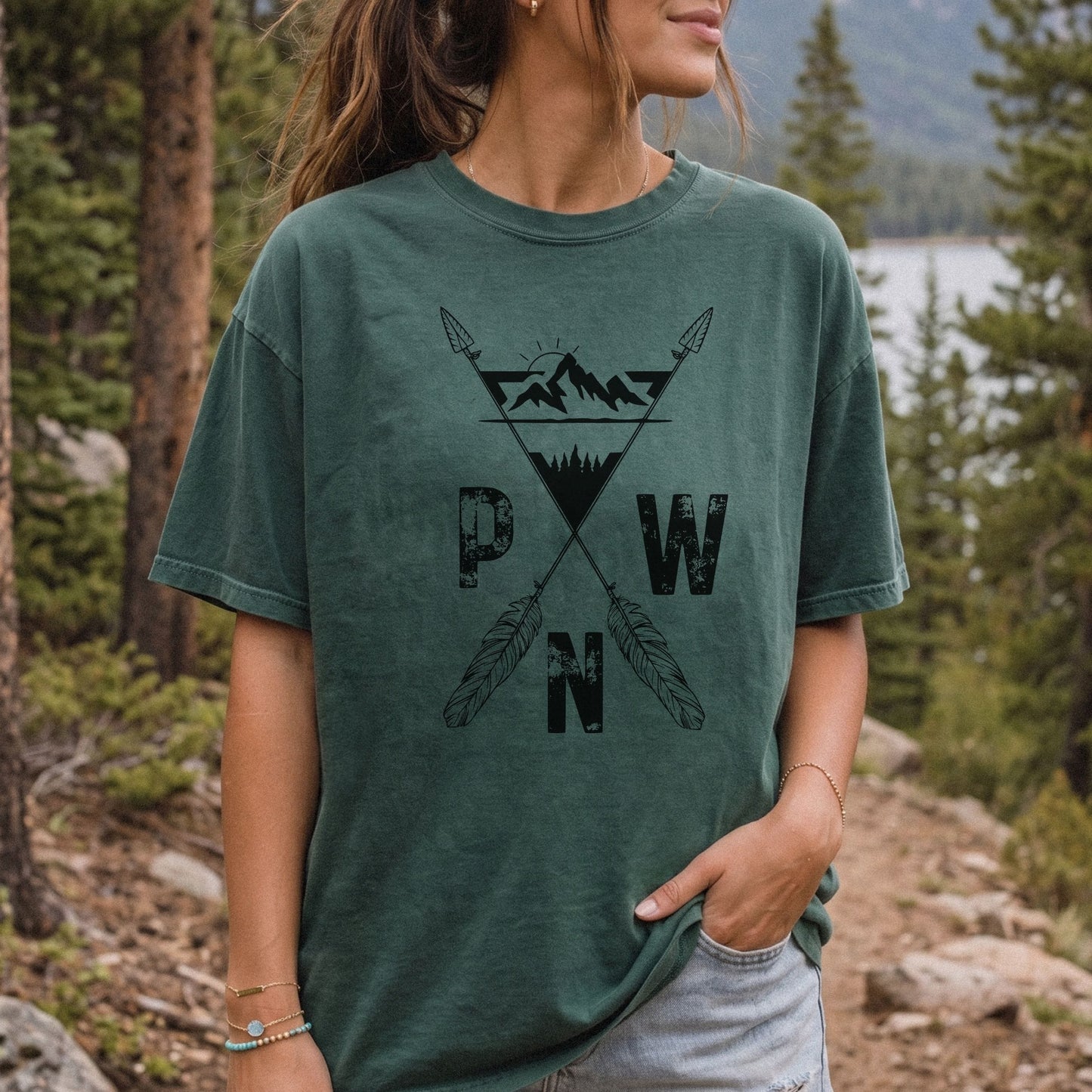 A woman wearing a dark green graphic t-shirt with a black mountain and arrow design spelling 'PWN' in an outdoor forest setting.
