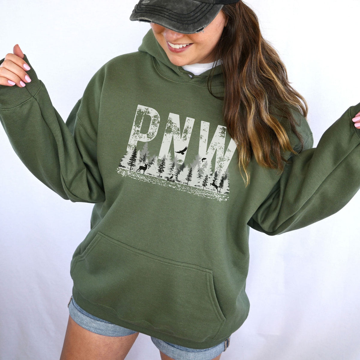 A woman wearing a military green pullover hoodie with a distressed white 'PNW' graphic featuring a forest and birds, paired with denim shorts and a black cap against a white studio background.