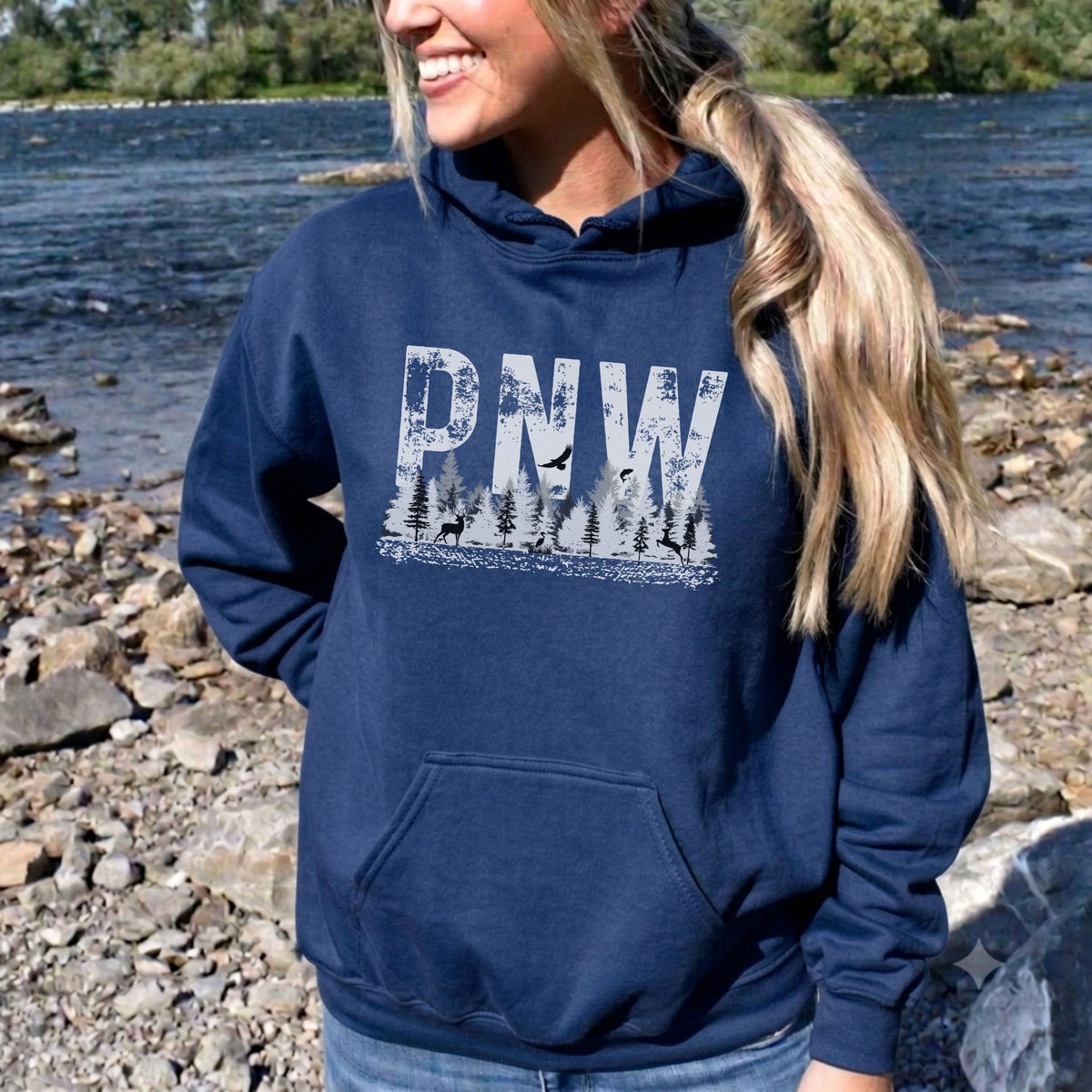 A woman wearing a navy blue hoodie with a distressed white 'PNW' graphic and pine tree silhouette, standing outdoors on a rocky riverbank.