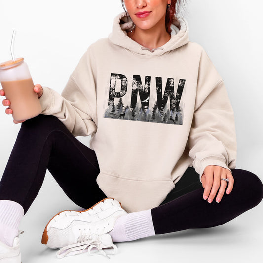 A woman sitting cross-legged wearing a beige pullover hoodie with a large black 'PNW' forest graphic on the chest, paired with black leggings and white sneakers. She is holding an iced coffee against a clean white background.