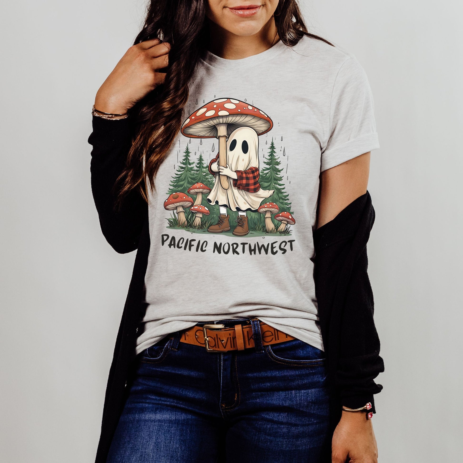 A woman wearing a light gray graphic t-shirt with a 'Pacific Northwest' mushroom and ghost design, styled with a black cardigan, brown belt, and blue jeans against a plain light gray background.