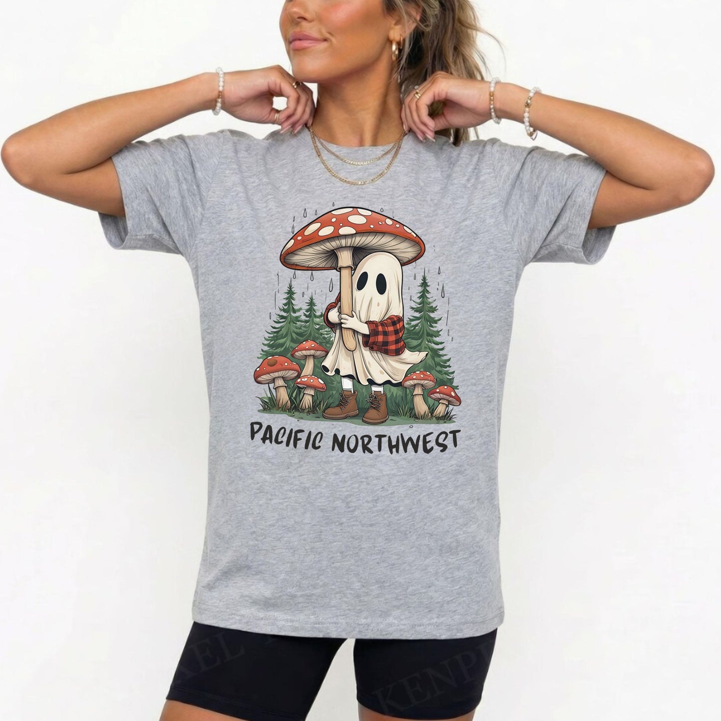 A woman wearing a heather gray graphic t-shirt with a cartoon ghost hugging a giant red and white mushroom in a forest, with the text 'PACIFIC NORTHWEST' printed below. She is also wearing black athletic shorts and gold jewelry against a plain white background.