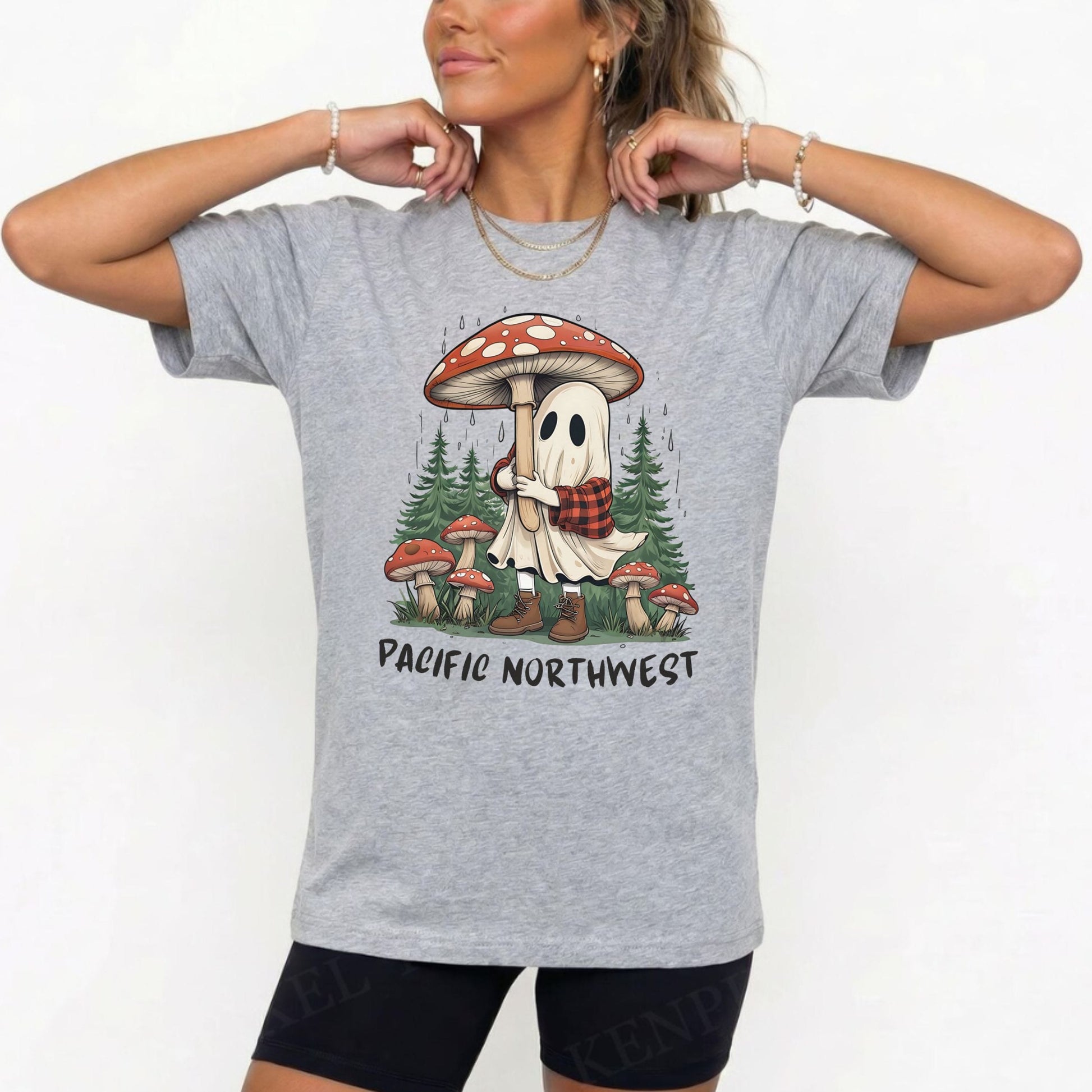 A woman wearing a heather gray graphic t-shirt with a cartoon ghost hugging a giant red and white mushroom in a forest, with the text 'PACIFIC NORTHWEST' printed below. She is also wearing black athletic shorts and gold jewelry against a plain white background.