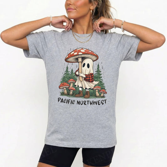A woman wearing a heather gray graphic t-shirt with a cartoon ghost hugging a giant red and white mushroom in a forest, with the text 'PACIFIC NORTHWEST' printed below. She is also wearing black athletic shorts and gold jewelry against a plain white background.