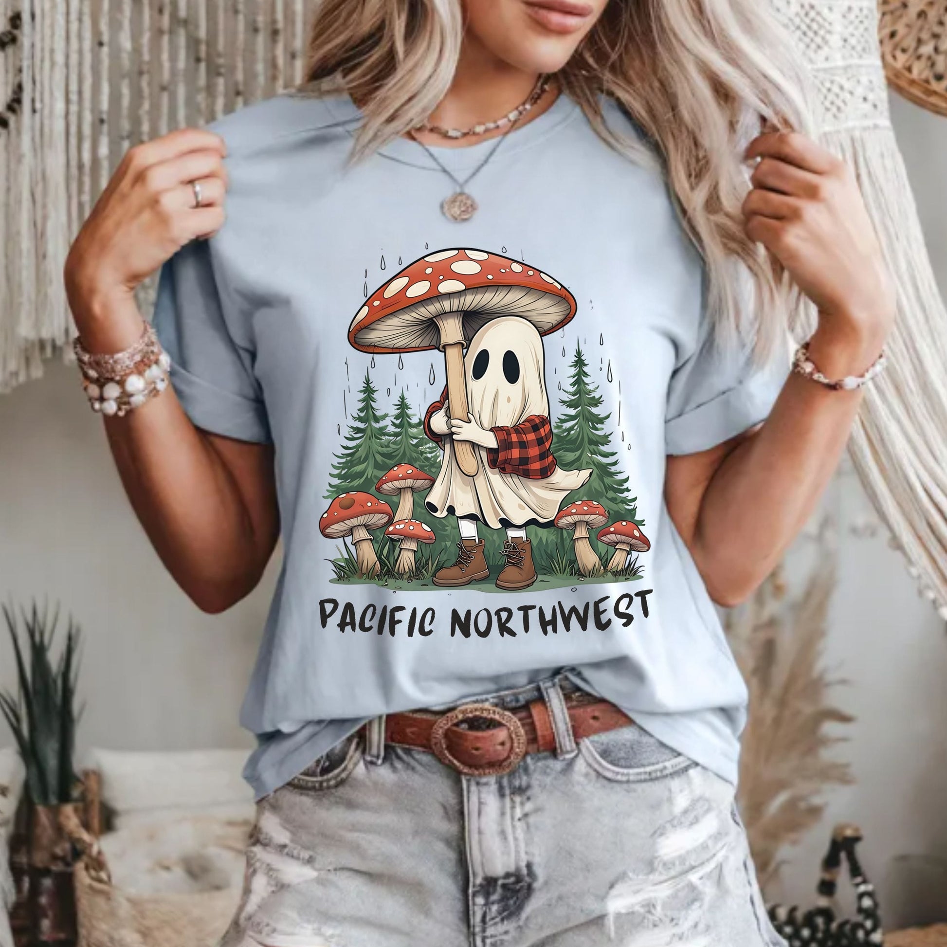 A woman wearing a light blue graphic t-shirt with a cartoon ghost holding a mushroom stick in a forest, with the text 'PACIFIC NORTHWEST' below. She is also wearing light-wash distressed jeans and a brown leather belt.
