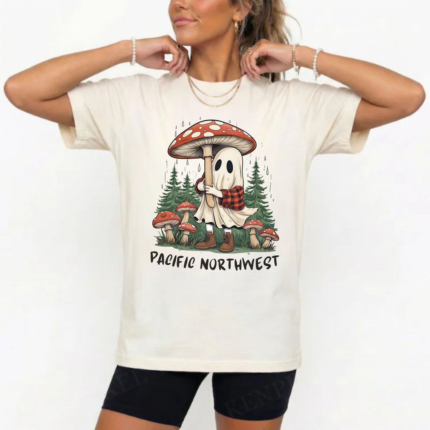 A woman wearing a cream-colored graphic t-shirt with a cartoon ghost and mushroom design, paired with black bike shorts, against a plain white background.