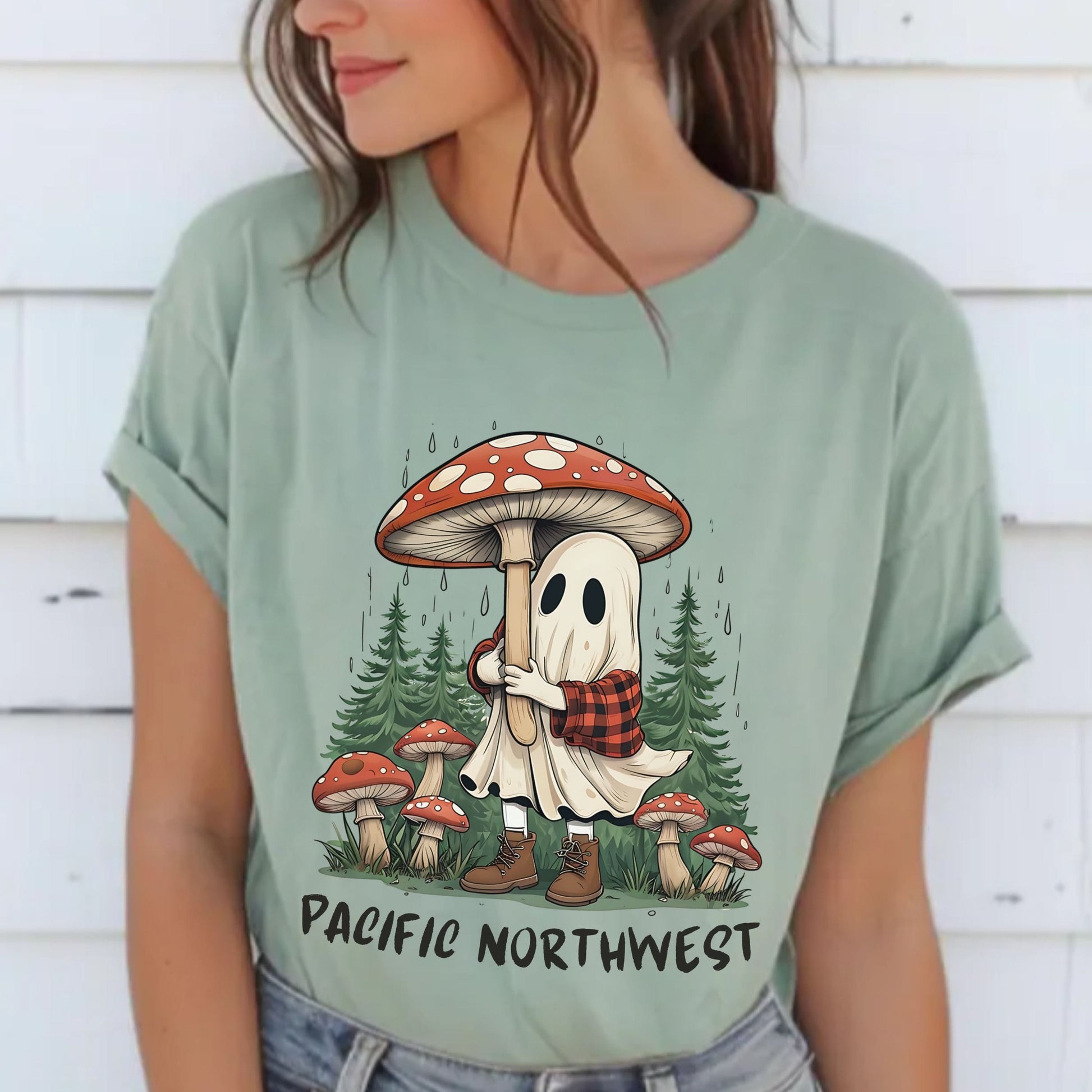A woman wearing a sage green graphic t-shirt with a cartoon ghost hugging a giant mushroom under pine trees, with the text 'PACIFIC NORTHWEST' printed below.