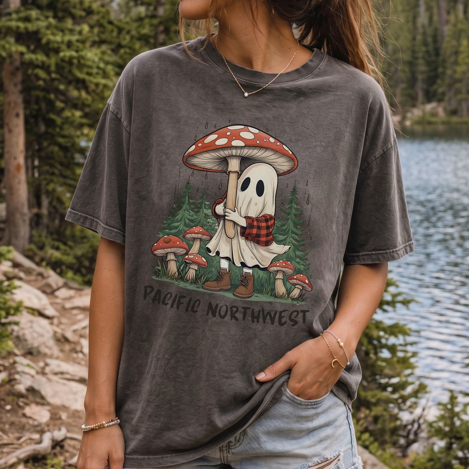 A woman wearing a dark gray mineral-wash graphic t-shirt featuring a cartoon ghost in a red and white mushroom cap holding a campfire, with 'PACIFIC NORTHWEST' text below, set against a background of a forest and lake.