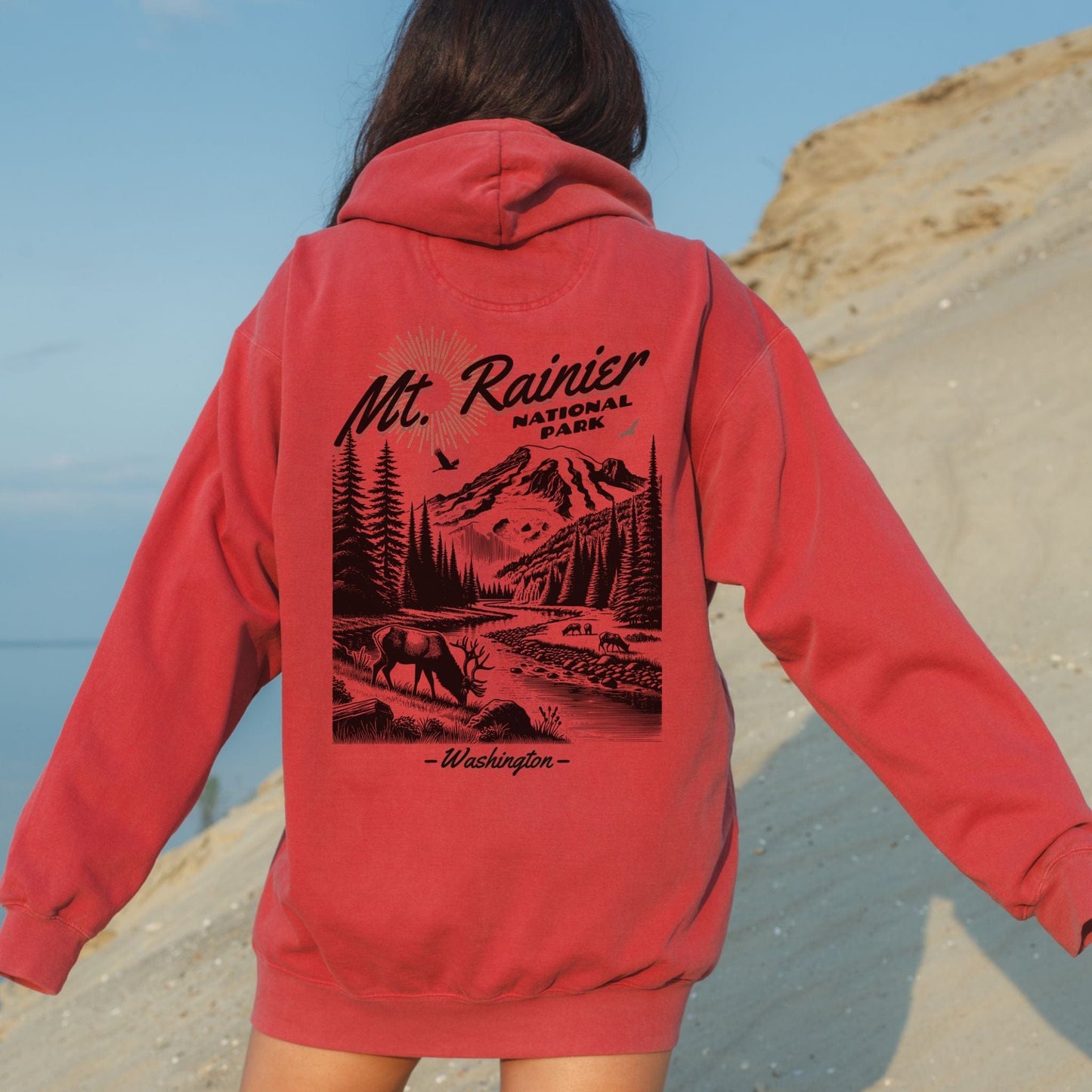 Person wearing a red hoodie with 'Mt. Rainier National Park, Washington' design on the back, standing on a beach.