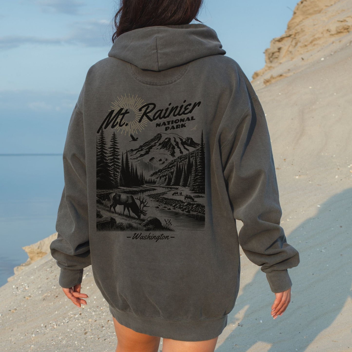 Back view of a woman on a sandy beach wearing a faded charcoal pepper Comfort Colors pullover hoodie featuring a black vintage illustration of an elk at Mt. Rainier National Park Washington.