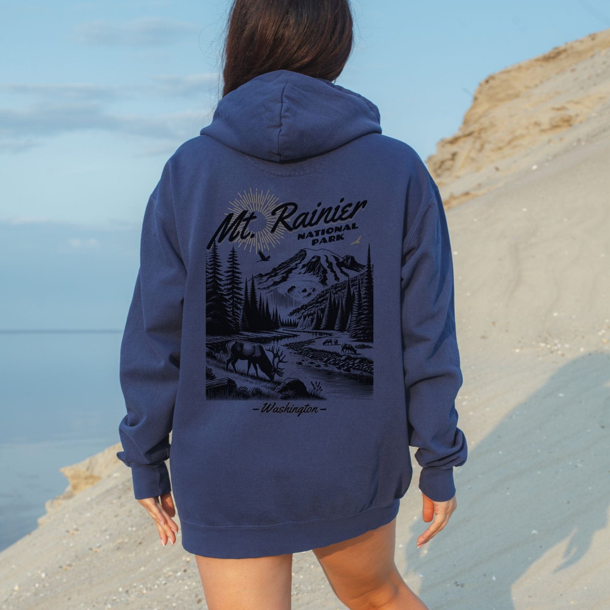 Person wearing a blue hoodie with Mount Rainier National Park design, standing on a beach.
