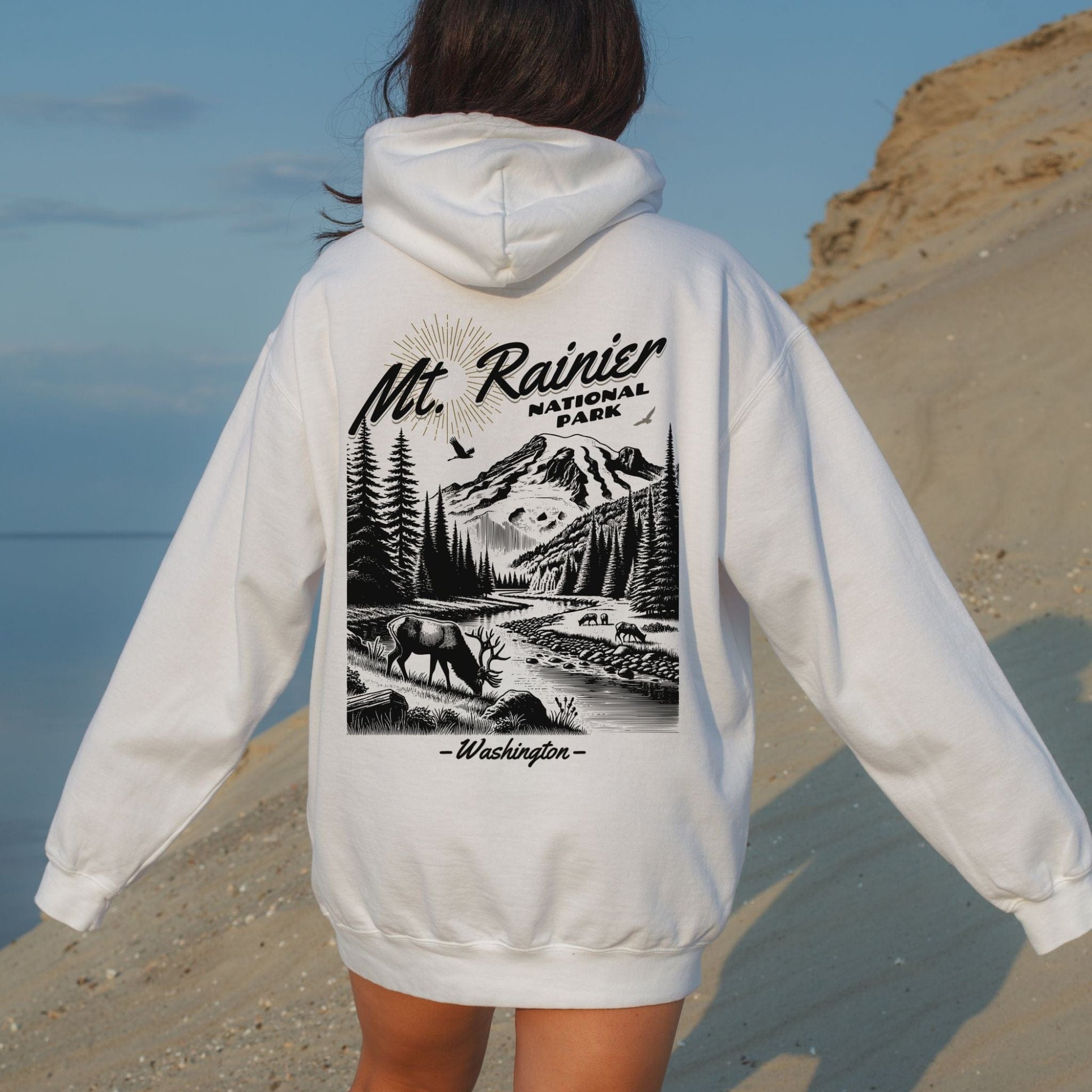 Back view of a woman on a sandy beach wearing a white Comfort Colors pullover hoodie featuring a black vintage illustration of an elk at Mt. Rainier National Park Washington.