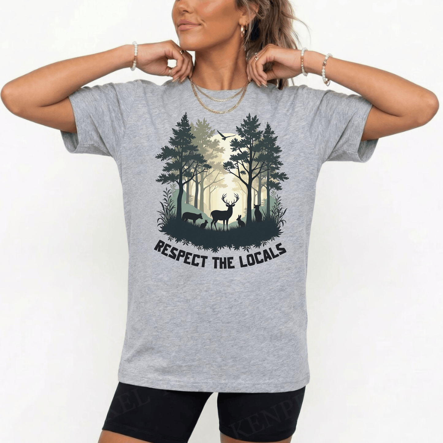 A woman wearing a heather gray graphic t-shirt with a forest design and the text 'RESPECT THE LOCALS'. The background is a clean white studio setting.