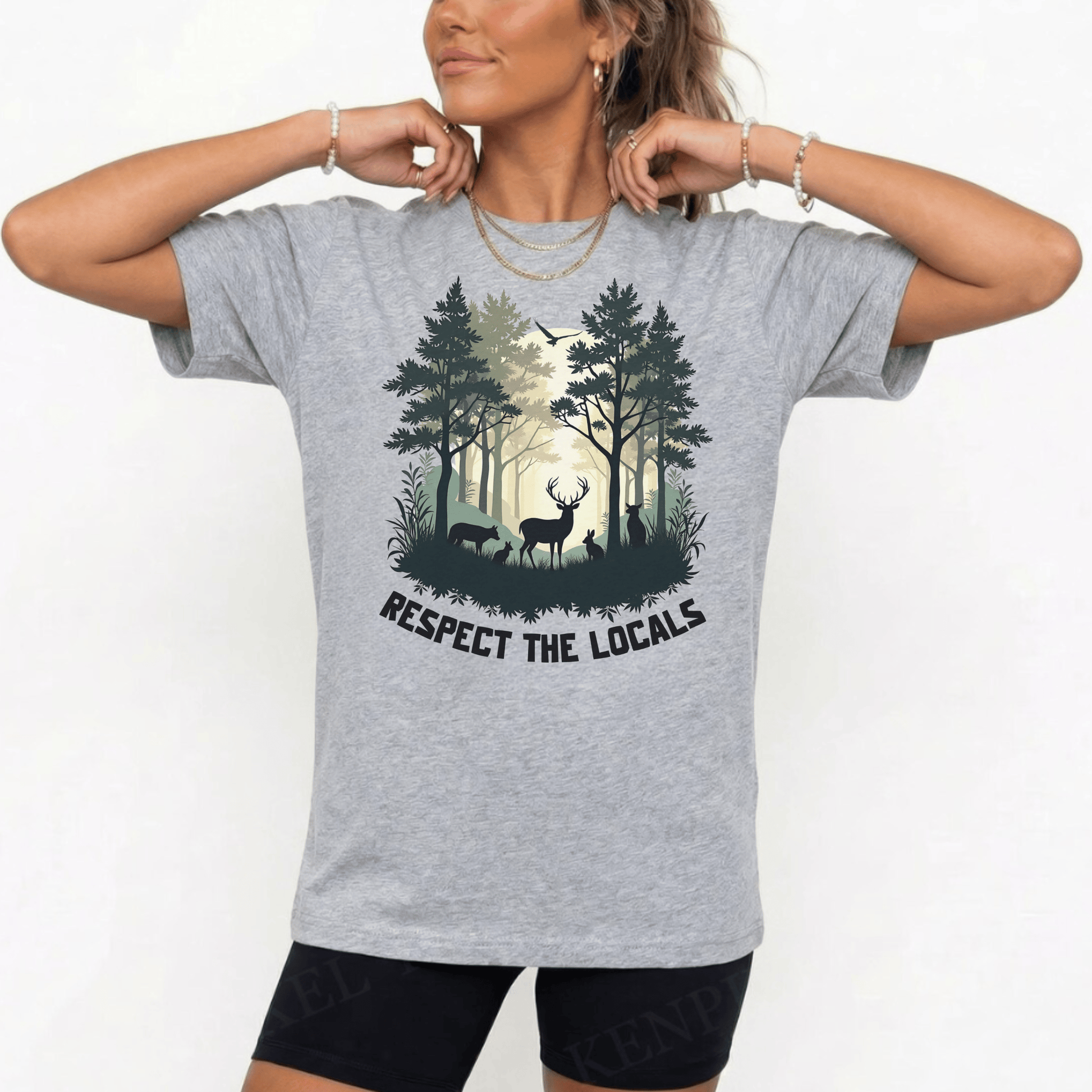 A woman wearing a heather gray graphic t-shirt with a forest design and the text 'RESPECT THE LOCALS'. The background is a clean white studio setting.