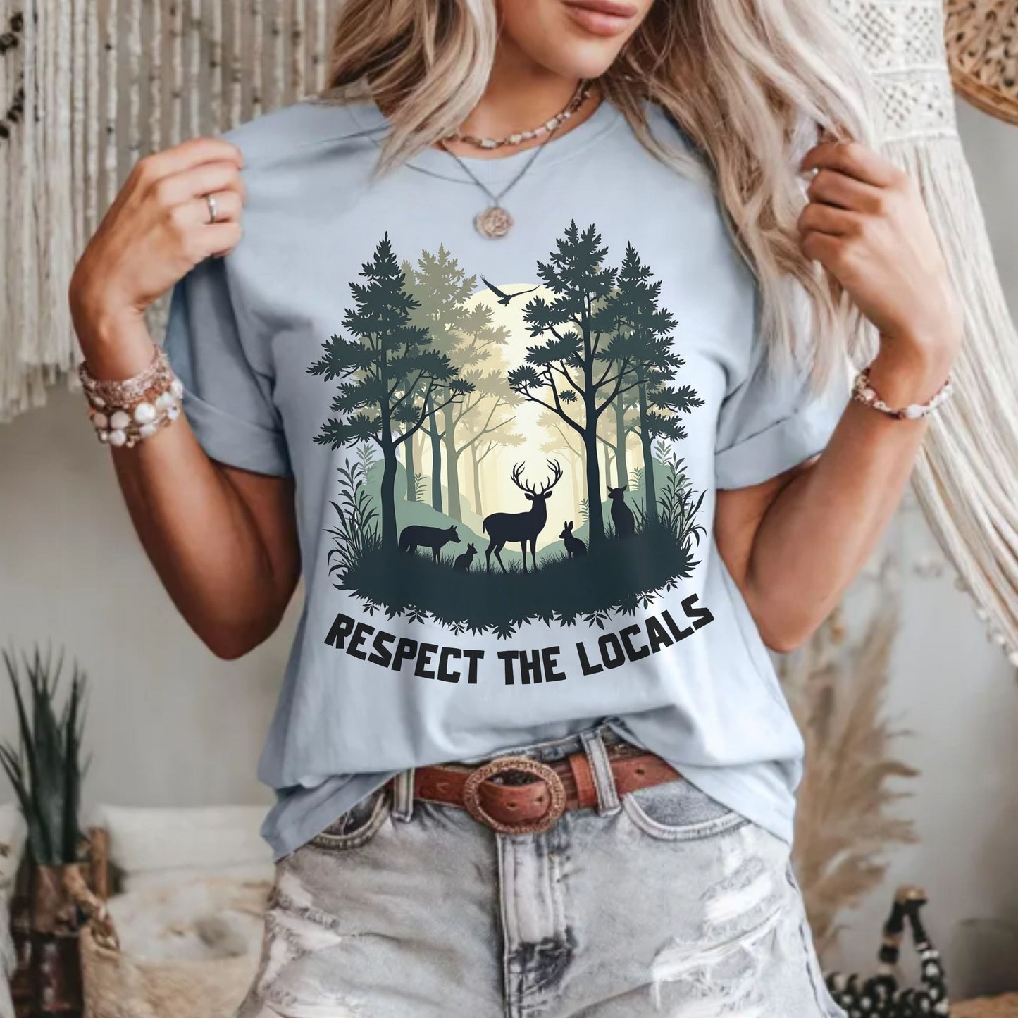 A woman wearing a light blue graphic t-shirt with a forest and deer design and the text 'RESPECT THE LOCALS'.