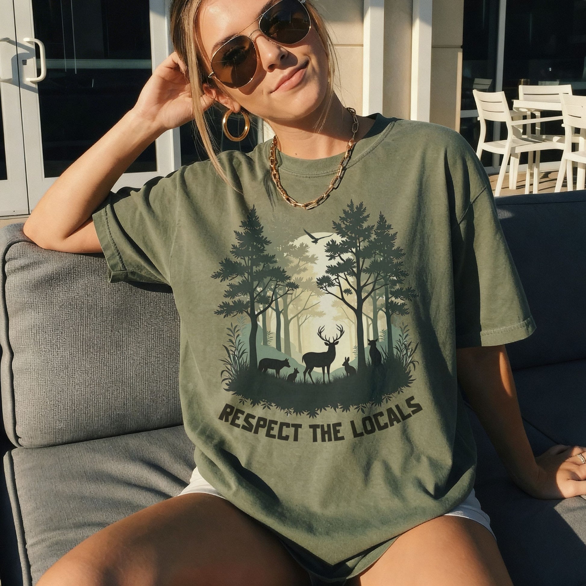 A woman wearing an oversized military green graphic t-shirt with a forest and deer design and the text 'RESPECT THE LOCALS', sitting on a gray outdoor sofa.