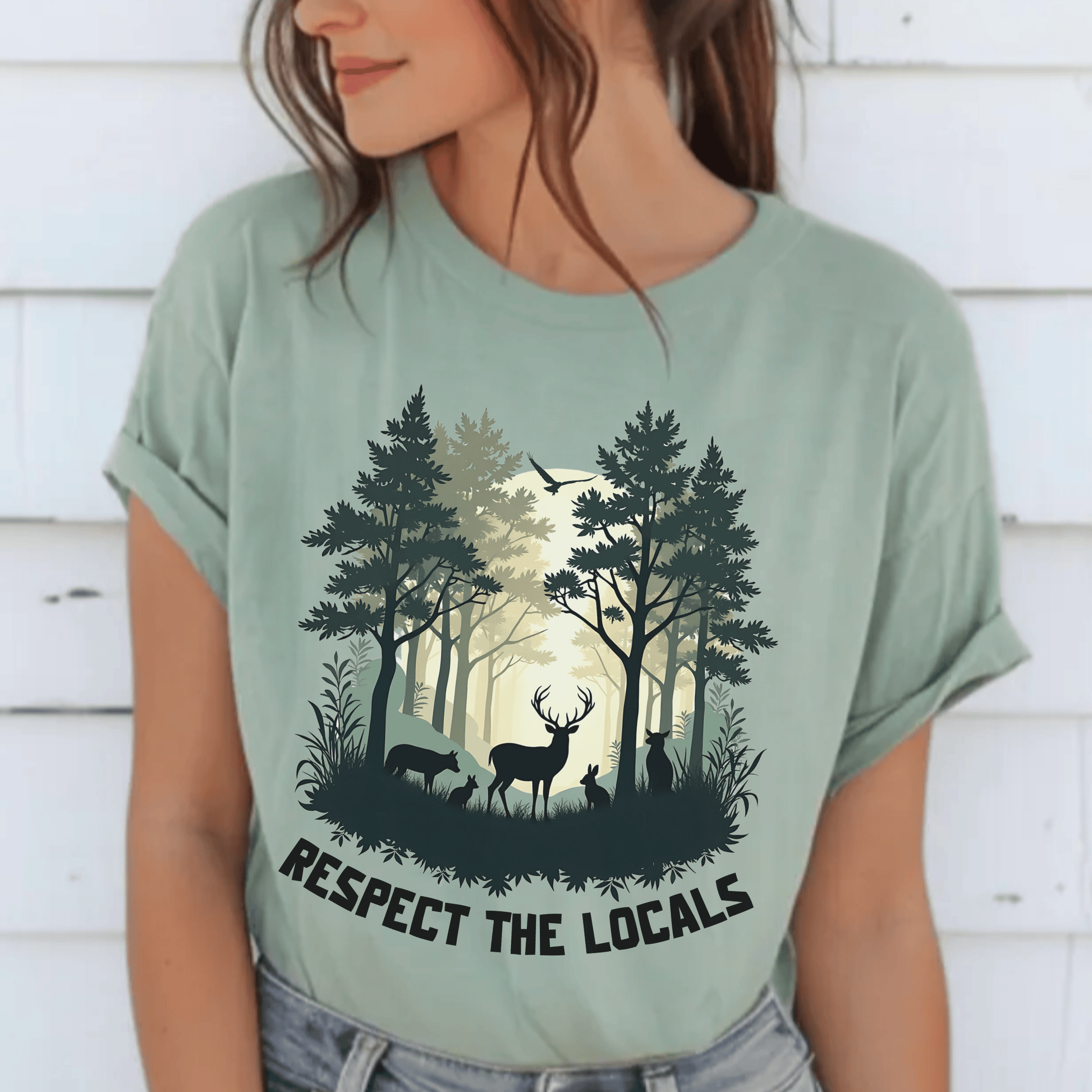 A woman wearing a sage green graphic t-shirt with a forest and deer design and the text 'RESPECT THE LOCALS'.