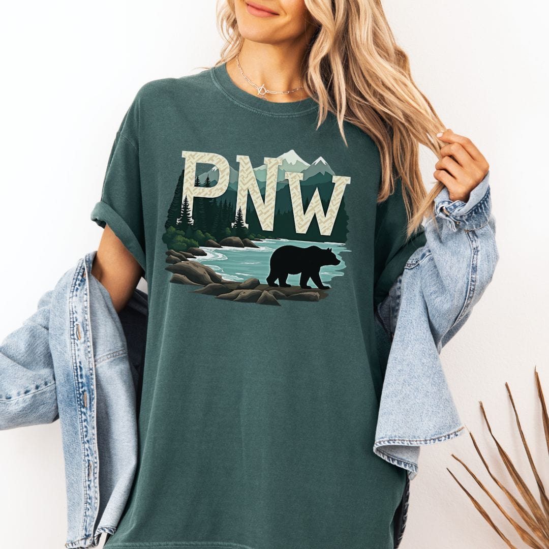 Woman wearing a green t-shirt with PNW graphic featuring a bear and mountains on a white background
