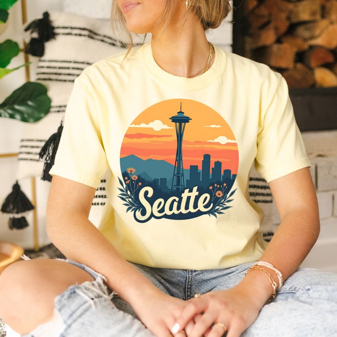 Woman wearing a light yellow graphic t-shirt featuring a retro circular print of the Seattle skyline and Space Needle against a vibrant orange and pink sunset with a botanical floral border.
