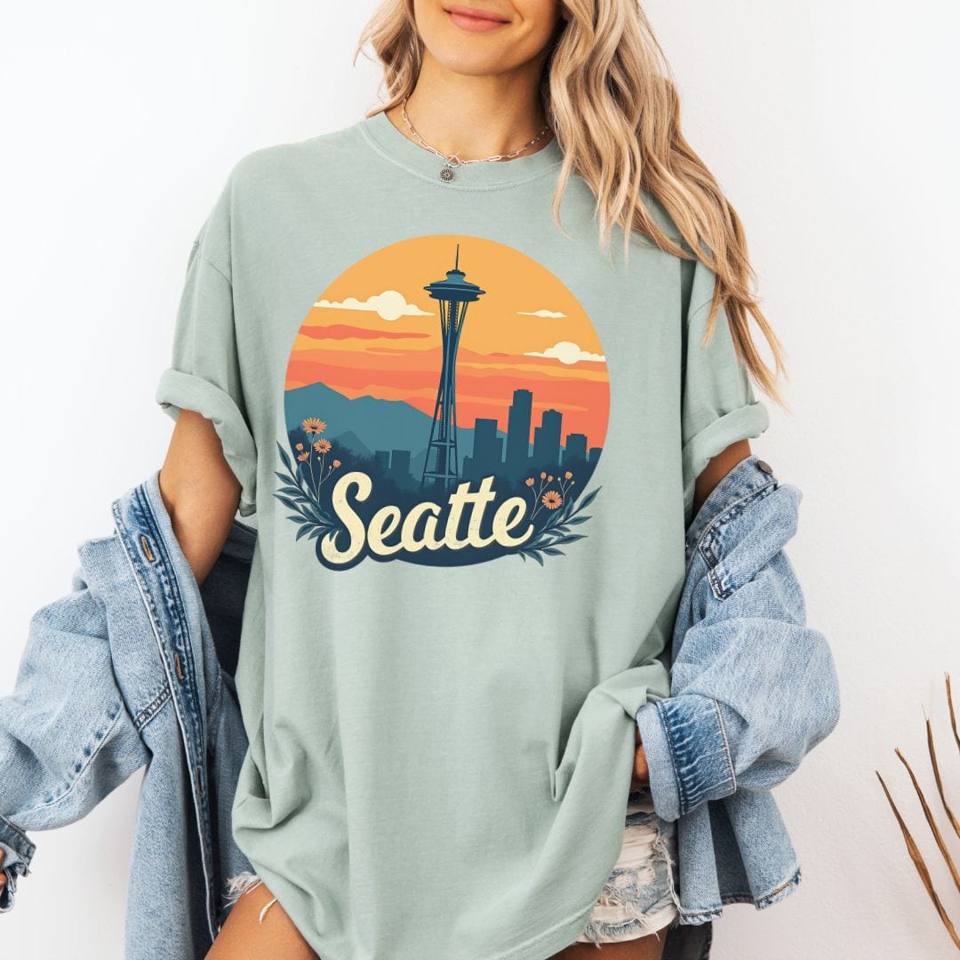 Woman wearing a sage green graphic t-shirt featuring a retro circular print of the Seattle skyline and Space Needle against a vibrant orange and pink sunset with a botanical floral border.