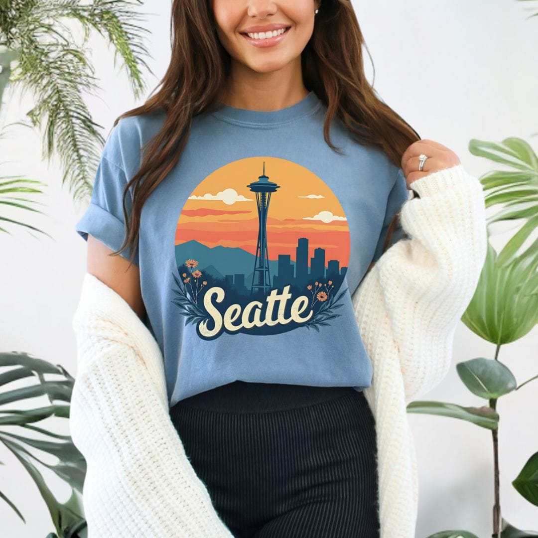Woman wearing a blue jean colored graphic t-shirt featuring a retro circular print of the Seattle skyline and Space Needle against a vibrant orange and pink sunset with a botanical floral border.