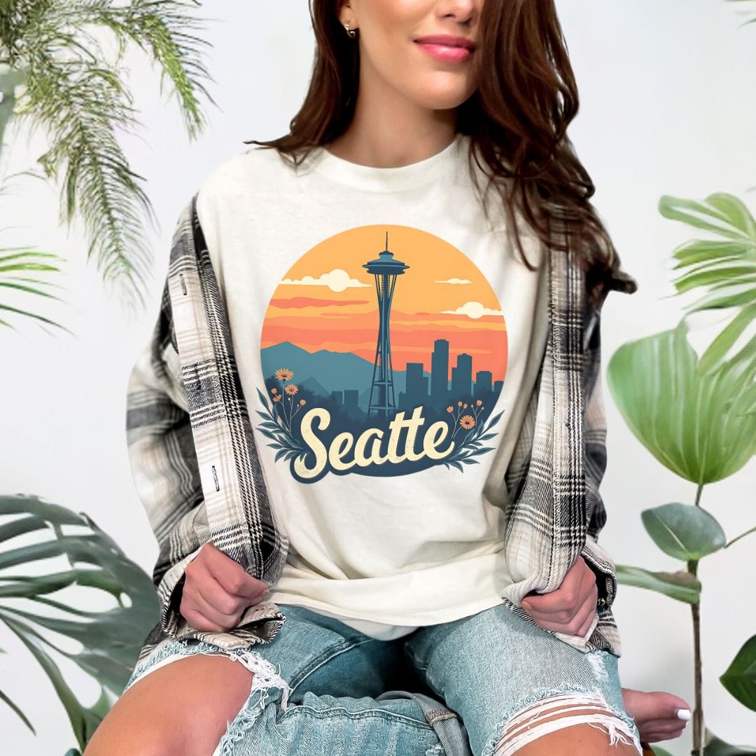 Woman wearing a cream colored graphic t-shirt featuring a retro circular print of the Seattle skyline and Space Needle against a vibrant orange and pink sunset with a botanical floral border.