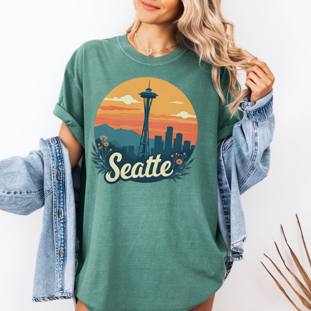Woman wearing a light yellow graphic t-shirt featuring a retro circular print of the Seattle skyline and Space Needle against a vibrant orange and pink sunset with a botanical floral border.