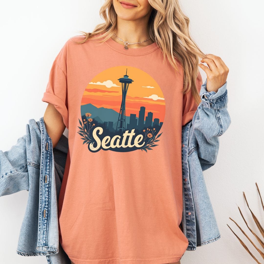 Woman wearing a terracotta colored graphic t-shirt featuring a retro circular print of the Seattle skyline and Space Needle against a vibrant orange and pink sunset with a botanical floral border.