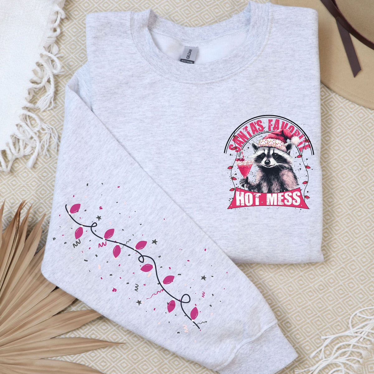 A gray sweatshirt with a raccoon design and the text 'Santa's Favorite Hot Mess' on the front, displayed on a flat surface with a beige hat and a decorative branch in the background.