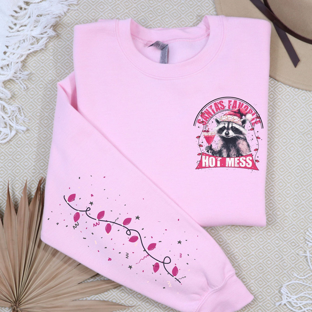 A pink sweatshirt with a raccoon design and the text 'Santa's Favorite Hot Mess' on the front, displayed on a flat surface with a beige hat and a decorative branch in the background.