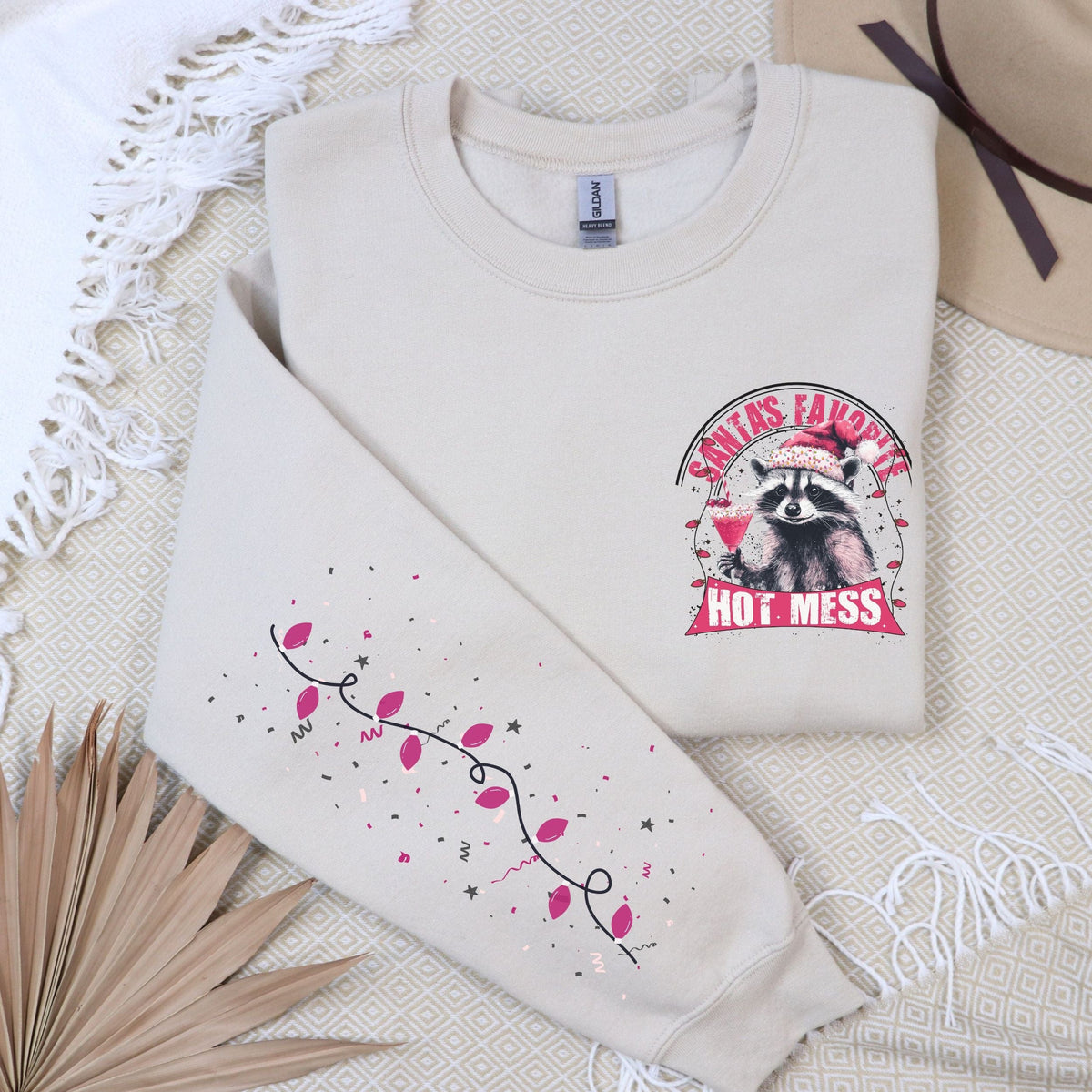 A beige sweatshirt with a festive raccoon design and the text 'Santa's Favorite Hot Mess' on the front, displayed on a flat surface with a beige hat and a decorative branch in the background.