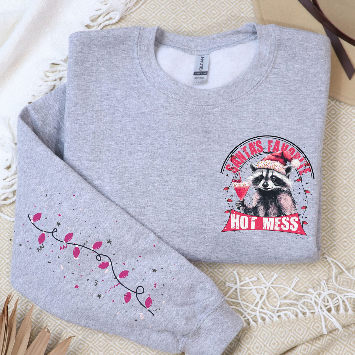 A gray sweatshirt with a festive raccoon design and the text 'Santa's Favorite Hot Mess' on the front, displayed on a flat surface with a beige hat and a decorative branch in the background.