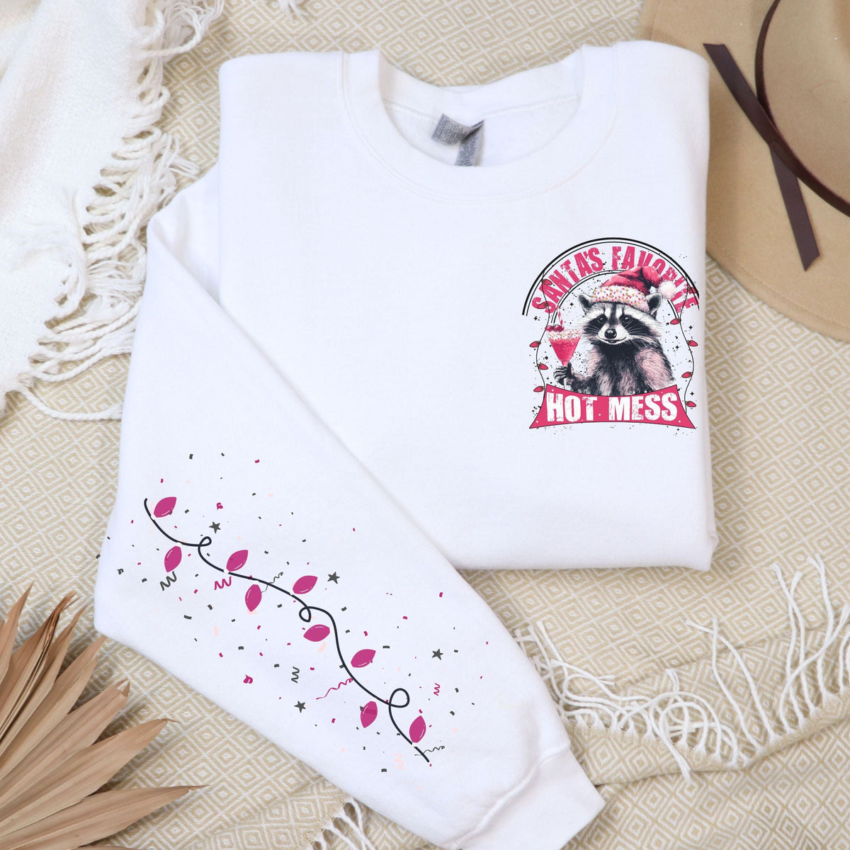 A shite sweatshirt with a holiday raccoon design and the text 'Santa's Favorite Hot Mess' on the front, displayed on a flat surface with a beige hat and a decorative branch in the background.