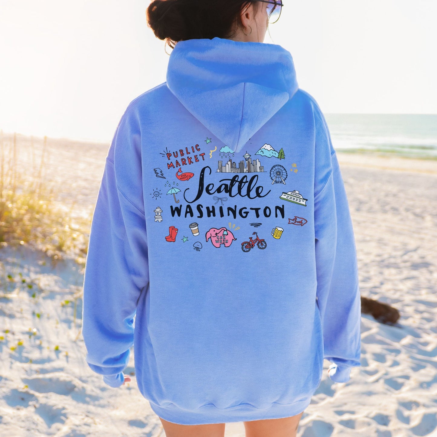 A person standing on a sandy beach wearing an oversized light blue hoodie, shown from the back. The hoodie features a colorful graphic print with the words 'Seattle Washington' and small illustrated city-themed details.